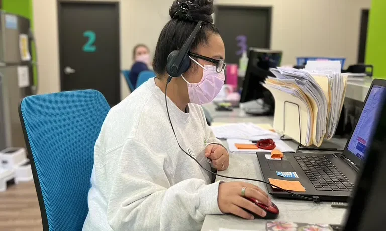 A woman wearing a pink mask and headset is sitting at a desk, working on a laptop. She has a stack of papers beside her and is concentrating on the screen. The office has colorful decor with green and blue accents.