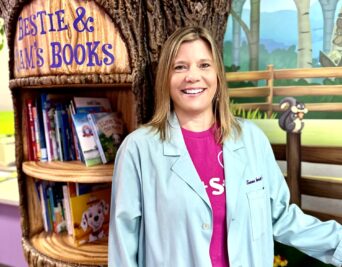 A smiling woman in a light blue lab coat stands near a whimsical bookshelf designed to resemble a tree. The shelf holds childrens books, and the backdrop features a vibrant, cartoon-like forest mural.