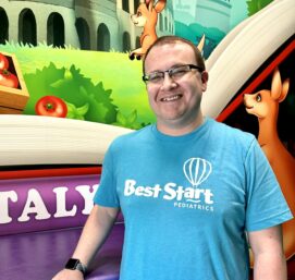 A smiling man wearing glasses and a blue Best Start Pediatrics shirt stands indoors. The colorful background features illustrations of kangaroos, tomatoes, and large books.