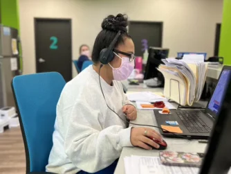 A woman wearing a pink mask and headset is sitting at a desk, working on a laptop. She has a stack of papers beside her and is concentrating on the screen. The office has colorful decor with green and blue accents.