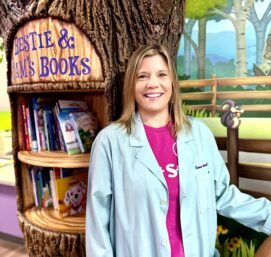 A smiling person in a light blue coat stands next to a tree-shaped bookshelf filled with childrens books. The background features a painted mural with trees and a fence, creating a playful and colorful setting.