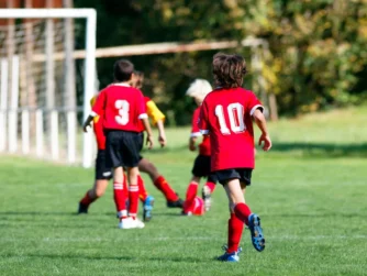 Four children playing soccer on a grassy field. Two are wearing red jerseys, one with the number 10 and another with number 3. They are running towards a goalpost in the background. Trees and a fence are visible in the distance.