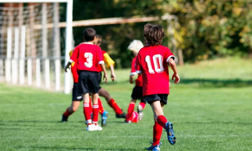 Four children playing soccer on a grassy field. Two are wearing red jerseys, one with the number 10 and another with number 3. They are running towards a goalpost in the background. Trees and a fence are visible in the distance.