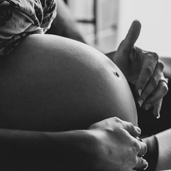 Black and white image of a pregnant belly with hands gently resting on it. The focus is on the roundness of the belly and the nurturing gesture of the hands, conveying a sense of anticipation and care.