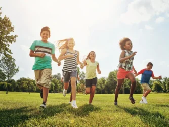 Five children are running joyfully across a sunny, grassy field. They are smiling and dressed in casual summer clothes. Trees and a bright sky fill the background, creating a cheerful and energetic scene.