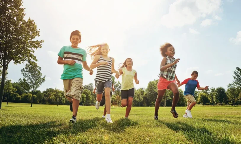 Five children are running joyfully across a sunny, grassy field. They are smiling and dressed in casual summer clothes. Trees and a bright sky fill the background, creating a cheerful and energetic scene.