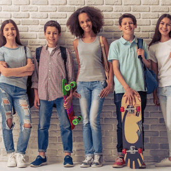 Six teenagers, four girls and two boys, stand in front of a white brick wall. They are casually dressed, smiling, and holding skateboards. They each wear different outfits, and some have backpacks. The mood is relaxed and cheerful.