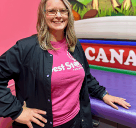 A woman with long hair and glasses, wearing a pink Best Start Pediatrics t-shirt and a dark jacket, smiles while standing in a colorful room with a mural of trees and the word CANADA visible behind her.