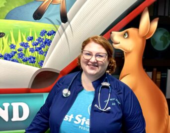 A woman wearing glasses and a stethoscope smiles while standing in front of a colorful mural featuring kangaroos. She is dressed in a blue jacket and a teal shirt with a visible logo. The mural depicts a bright, animated environment.