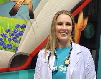 A person wearing a white coat and a teal shirt with a stethoscope around their neck smiles at the camera. They stand in front of a colorful, cartoonish wall with illustrations of flowers and animals.