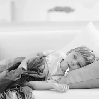 A young child lying on a couch with a blanket, holding a thermometer in their mouth, looking slightly unwell. The black and white image captures a calm, cozy indoor setting.