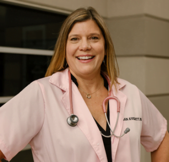 A smiling woman wearing a light pink lab coat and a stethoscope around her neck stands in front of a building. She has long, light brown hair and is posing with her hands on her hips.