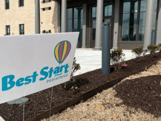 Sign reading Best Start Pediatrics with a colorful balloon logo in front of a two-story building. The building has large glass windows and multiple columns, with a small landscaped area in front.