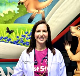 A smiling person stands in front of a colorful mural featuring kangaroos and flowers. They are wearing a white coat over a pink shirt with a stethoscope around their neck.