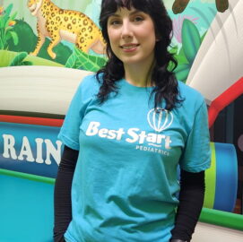 A person with dark hair is smiling and wearing a blue Best Start Pediatrics t-shirt. They are standing in front of a colorful wall mural featuring cartoon animals and jungle scenery.