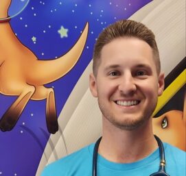 A person with short hair smiles while wearing a blue Best Start Pediatrics t-shirt and a stethoscope around their neck. The background features a colorful, cartoon-like illustration of animals and stars.