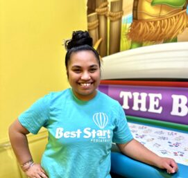 A person wearing a turquoise Best Start Pediatrics shirt smiles while leaning on a table in a vibrant room. The background features colorful wall art depicting a turtle by the beach.