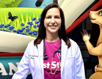 A woman in a white coat with a stethoscope stands smiling in front of a colorful mural with kangaroos, greenery, and a butterfly.