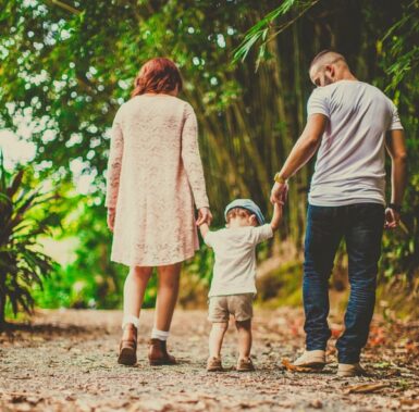 A family walks hand in hand down a leaf-covered pathway surrounded by lush greenery. The child is in the middle, wearing a light blue hat and shorts, while the adults wear casual clothes. The scene conveys a sense of togetherness and nature.
