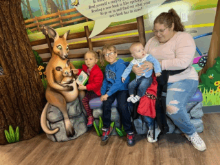 A woman and four children sit on a bench beside a kangaroo statue holding a book. The colorful background features a tree and a wooden fence. The children are dressed in casual clothing, two wearing glasses. They appear to be in a playful environment.
