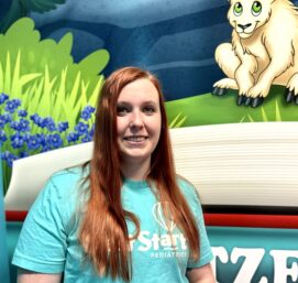 A person with long red hair, wearing a light blue shirt with text, stands in front of a colorful mural featuring a cartoon goat and plants.