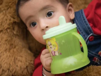 A baby with big eyes, wearing a red shirt and blue overalls, is lying on a soft brown surface, holding a green sippy cup with cartoon characters on it. The baby is gazing intently while gripping the cup.