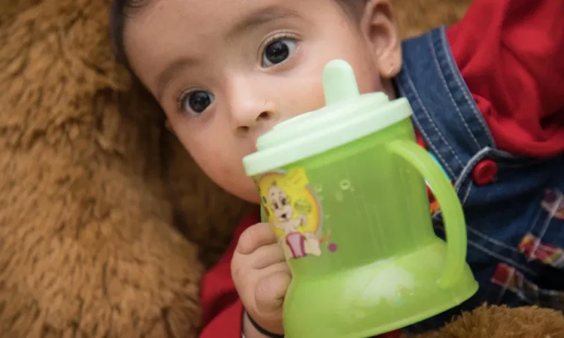 A baby with big eyes, wearing a red shirt and blue overalls, is lying on a soft brown surface, holding a green sippy cup with cartoon characters on it. The baby is gazing intently while gripping the cup.