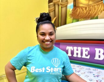 A smiling person wearing a teal Best Start Pediatrics shirt stands next to a colorful medical examination table, with a mural of a turtle and a beach scene in the background.