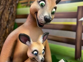 A cartoon kangaroo and joey sit together. The kangaroo is holding a book titled Cams Best Adventures. They are in front of a tree and a wooden fence, with a butterfly flying nearby. The setting is bright and colorful.