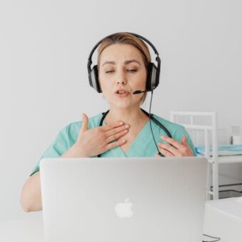 A healthcare professional wearing a headset conducts a virtual consultation on a laptop. She gestures towards her throat, possibly explaining or discussing a medical condition. She is dressed in a turquoise scrub uniform in a clinical setting.