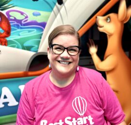 A person wearing glasses and a pink Best Start Pediatrics t-shirt smiling in front of a colorful mural with animals, including a kangaroo.