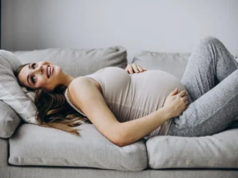 A pregnant woman lies on a couch, smiling and resting with a hand on her belly. She wears a light tank top and gray sweatpants, and her head is on a pillow. The background is a neutral-toned wall.