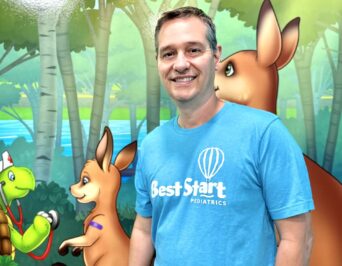 A smiling man in a blue Best Start Pediatrics shirt stands in front of a colorful mural featuring cartoon animals, including kangaroos and a turtle, amidst a forest scene.