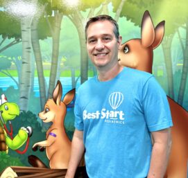 A man in a blue Best Start Pediatrics T-shirt stands smiling in front of a colorful mural featuring cartoon animals, including a turtle, two kangaroos, and a bird, set in a forest with trees and a river in the background.