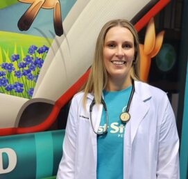 A person with long hair, wearing a white medical coat and a stethoscope, stands smiling in front of a colorful, cartoon-themed backdrop with flowers and animals.