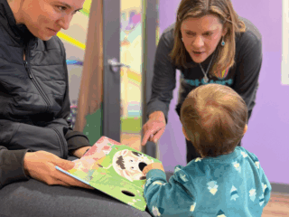 A woman sitting on a bench reads a picture book to a toddler standing close by. Another woman leans in, engaging with the child. The room has purple walls with colorful murals.